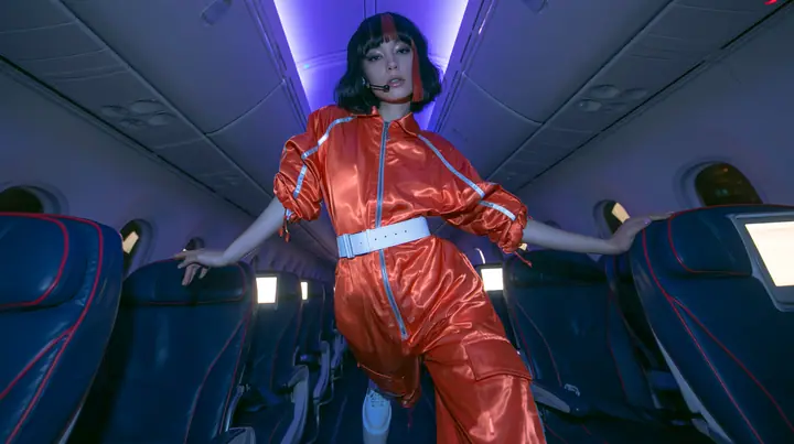 Jet-set high fashion editorial, a couture magazine story shot on a night flight: a full-length shot down the aisle of a Caucasian American woman in her late twenties with pale skin and a glossy black blunt bob with…