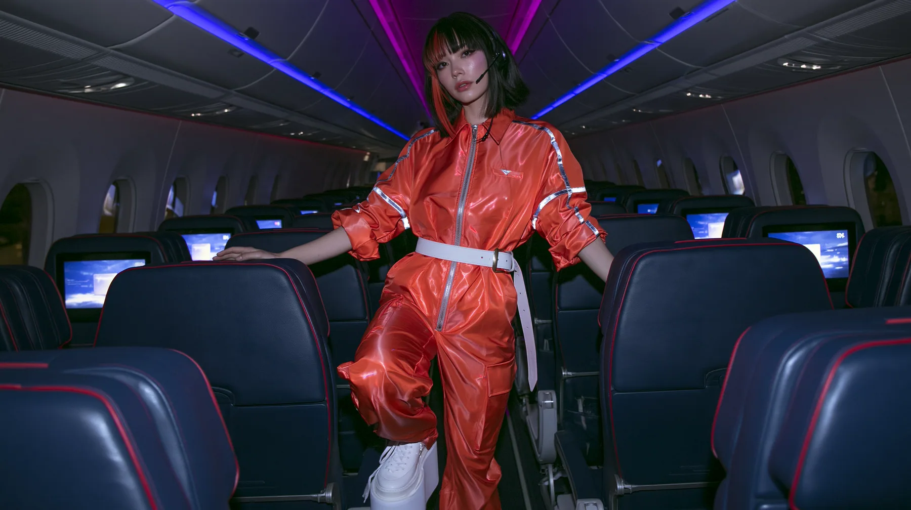 Jet-set high fashion editorial, a couture magazine story shot on a night flight: a full-length shot down the aisle of a Caucasian American woman in her late twenties with pale skin and a glossy black blunt bob with…