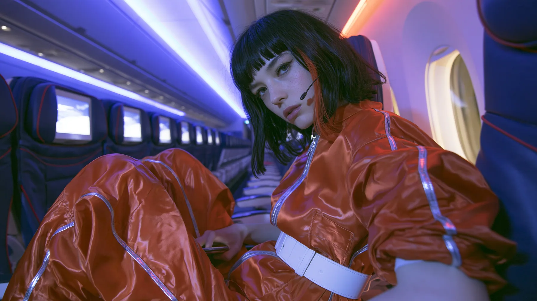 Jet-set high fashion editorial, a couture magazine story shot on a night flight: a medium-wide shot from the aisle of a Caucasian American woman in her late twenties with pale skin and a glossy black blunt bob with…