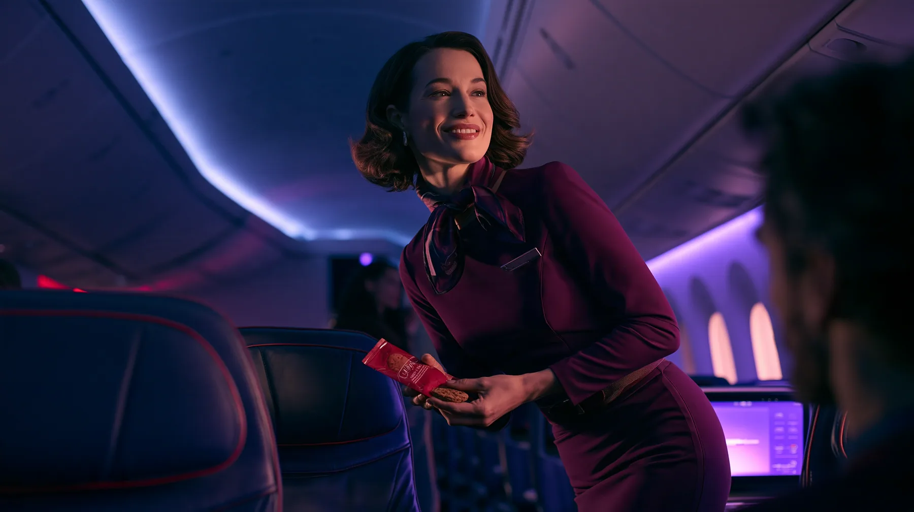 Jet-set high fashion editorial, a couture magazine story shot on a night flight: over the shoulder of a passenger in a window seat, an elegant flight attendant in her forties in a fitted plum-coloured dress and a…