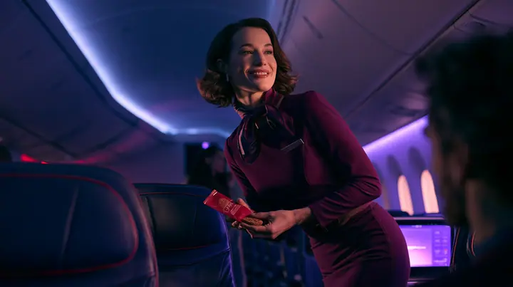 Jet-set high fashion editorial, a couture magazine story shot on a night flight: over the shoulder of a passenger in a window seat, an elegant flight attendant in her forties in a fitted plum-coloured dress and a…