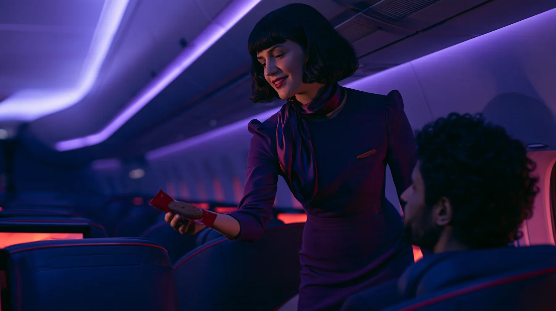 Jet-set high fashion editorial, a couture magazine story shot on a night flight: over the shoulder of a passenger in a window seat, an elegant flight attendant in her forties in a fitted plum-coloured dress and a…