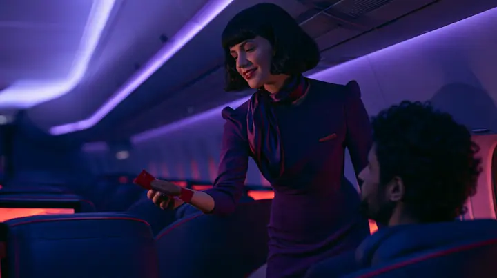 Jet-set high fashion editorial, a couture magazine story shot on a night flight: over the shoulder of a passenger in a window seat, an elegant flight attendant in her forties in a fitted plum-coloured dress and a…