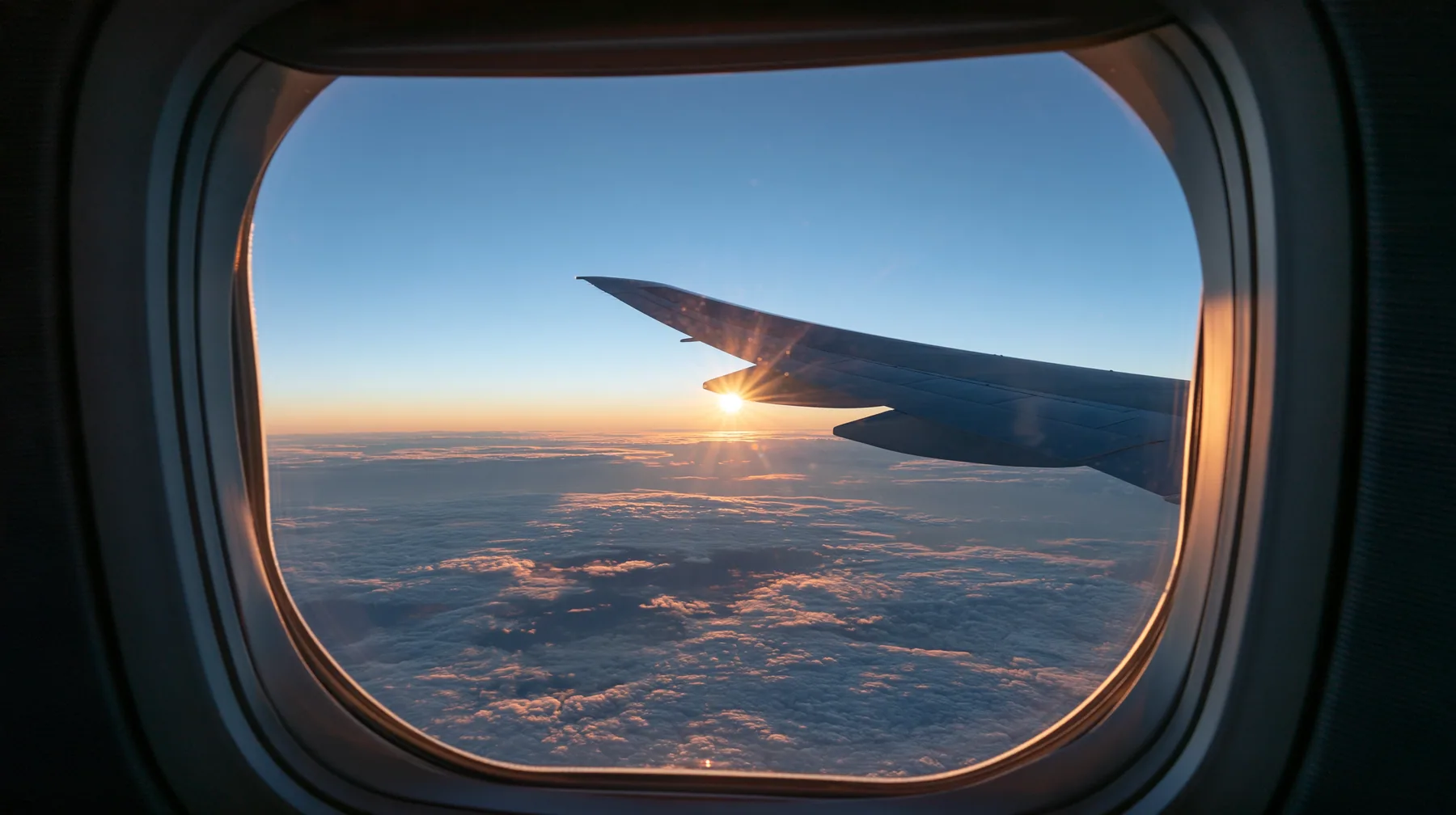 Jet-set editorial: the view out of an oval airliner window at dawn, the wing over a sea of clouds, the sun rising gold on the horizon, the window frame and shade in the foreground, 35mm lens, no text, no letters, no…