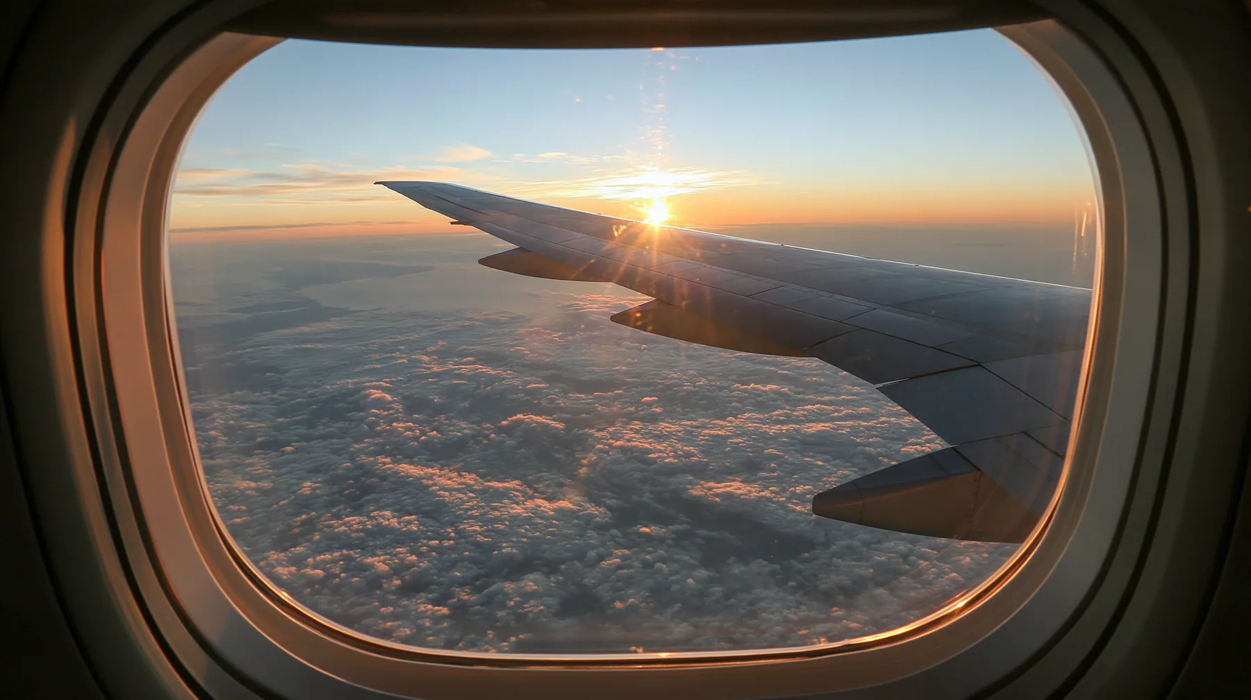 Jet-set editorial: the view out of an oval airliner window at dawn, the wing over a sea of clouds, the sun rising gold on the horizon, the window frame and shade in the foreground, 35mm lens, no text, no letters, no…
