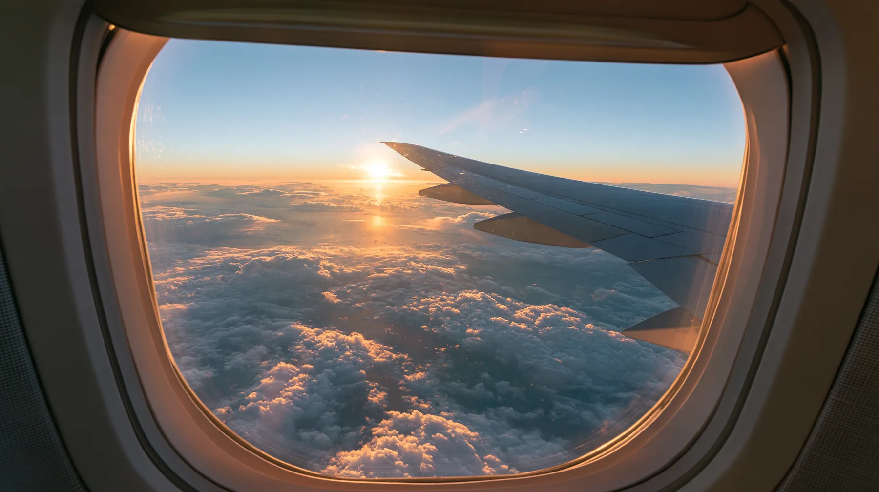 Jet-set editorial: the view out of an oval airliner window at dawn, the wing over a sea of clouds, the sun rising gold on the horizon, the window frame and shade in the foreground, 35mm lens, no text, no letters, no…
