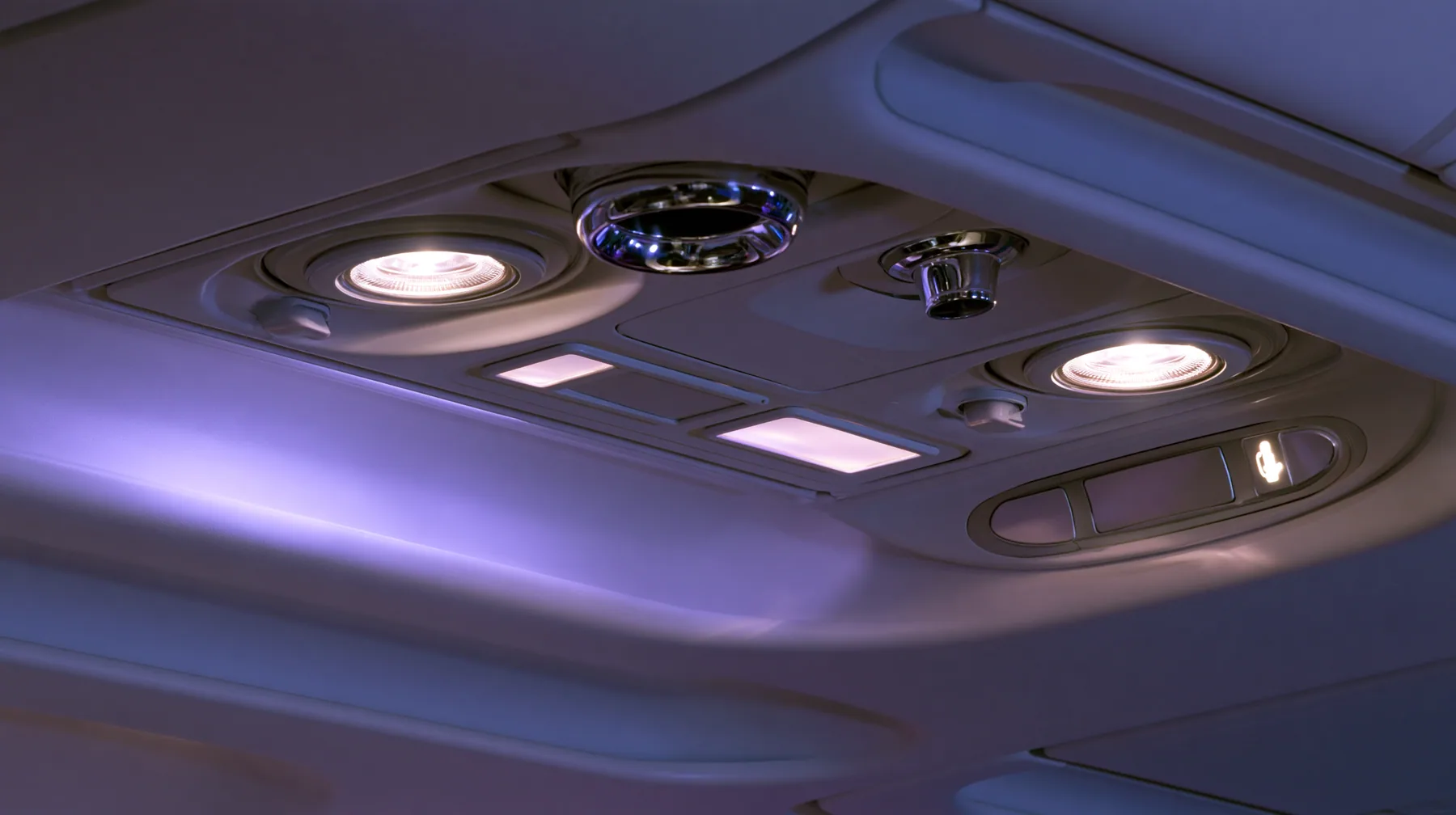 Jet-set editorial still life: an extreme close-up of the overhead passenger service panel above an airliner seat at night, two round reading lights, a chrome air nozzle, two small blank glowing sign panels, a…