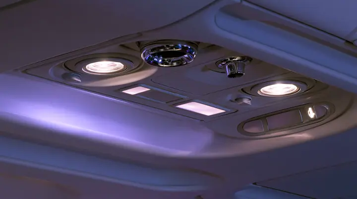 Jet-set editorial still life: an extreme close-up of the overhead passenger service panel above an airliner seat at night, two round reading lights, a chrome air nozzle, two small blank glowing sign panels, a…