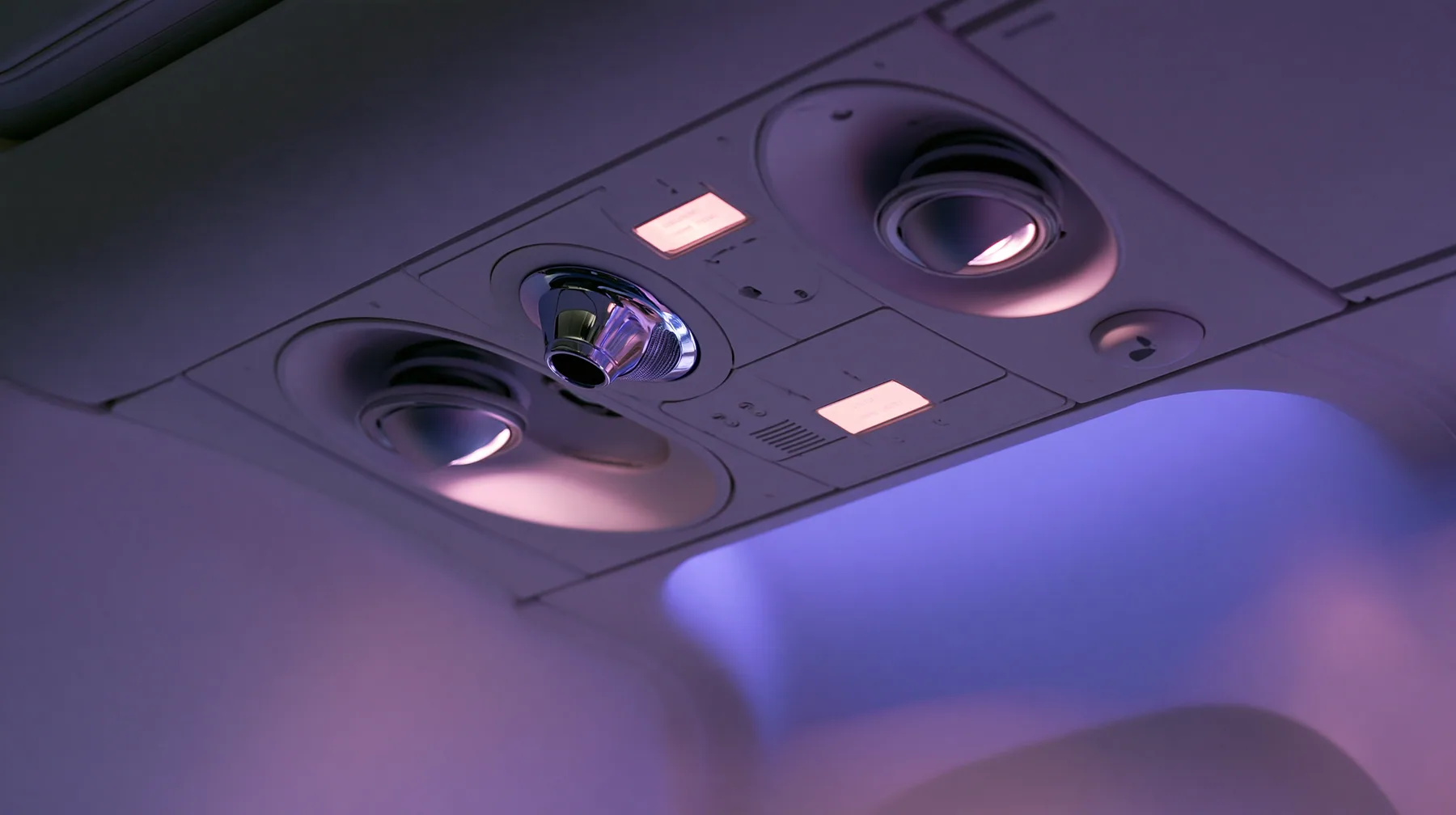 Jet-set editorial still life: an extreme close-up of the overhead passenger service panel above an airliner seat at night, two round reading lights, a chrome air nozzle, two small blank glowing sign panels, a…
