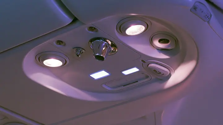 Jet-set editorial still life: an extreme close-up of the overhead passenger service panel above an airliner seat at night, two round reading lights, a chrome air nozzle, two small blank glowing sign panels, a…