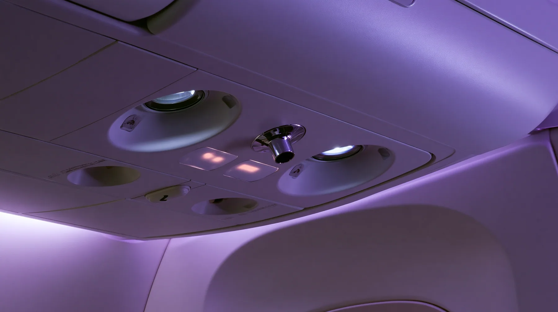 Jet-set editorial still life: an extreme close-up of the overhead passenger service panel above an airliner seat at night, two round reading lights, a chrome air nozzle, two small blank glowing sign panels, a…