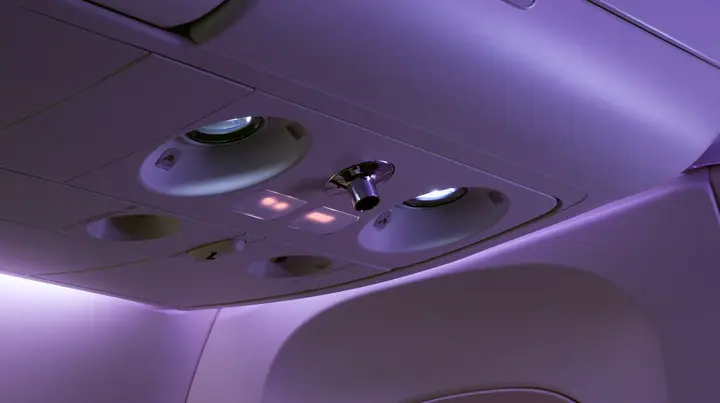Jet-set editorial still life: an extreme close-up of the overhead passenger service panel above an airliner seat at night, two round reading lights, a chrome air nozzle, two small blank glowing sign panels, a…