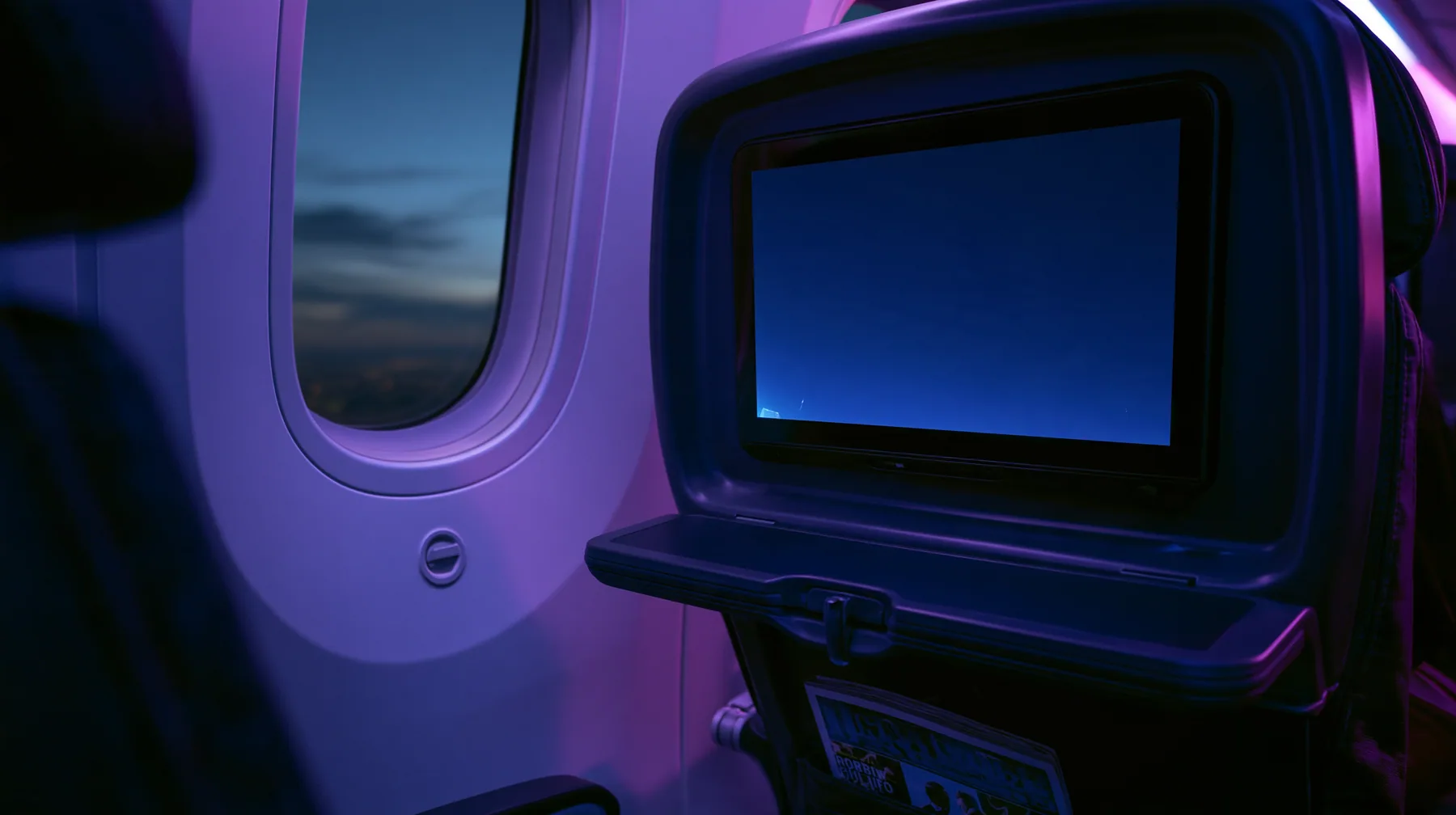 Jet-set editorial still life: the point of view from a window seat on a night flight, the seat-back screen in front glowing a blank deep navy, the tray table latched, a magazine in the seat pocket, the oval window at…