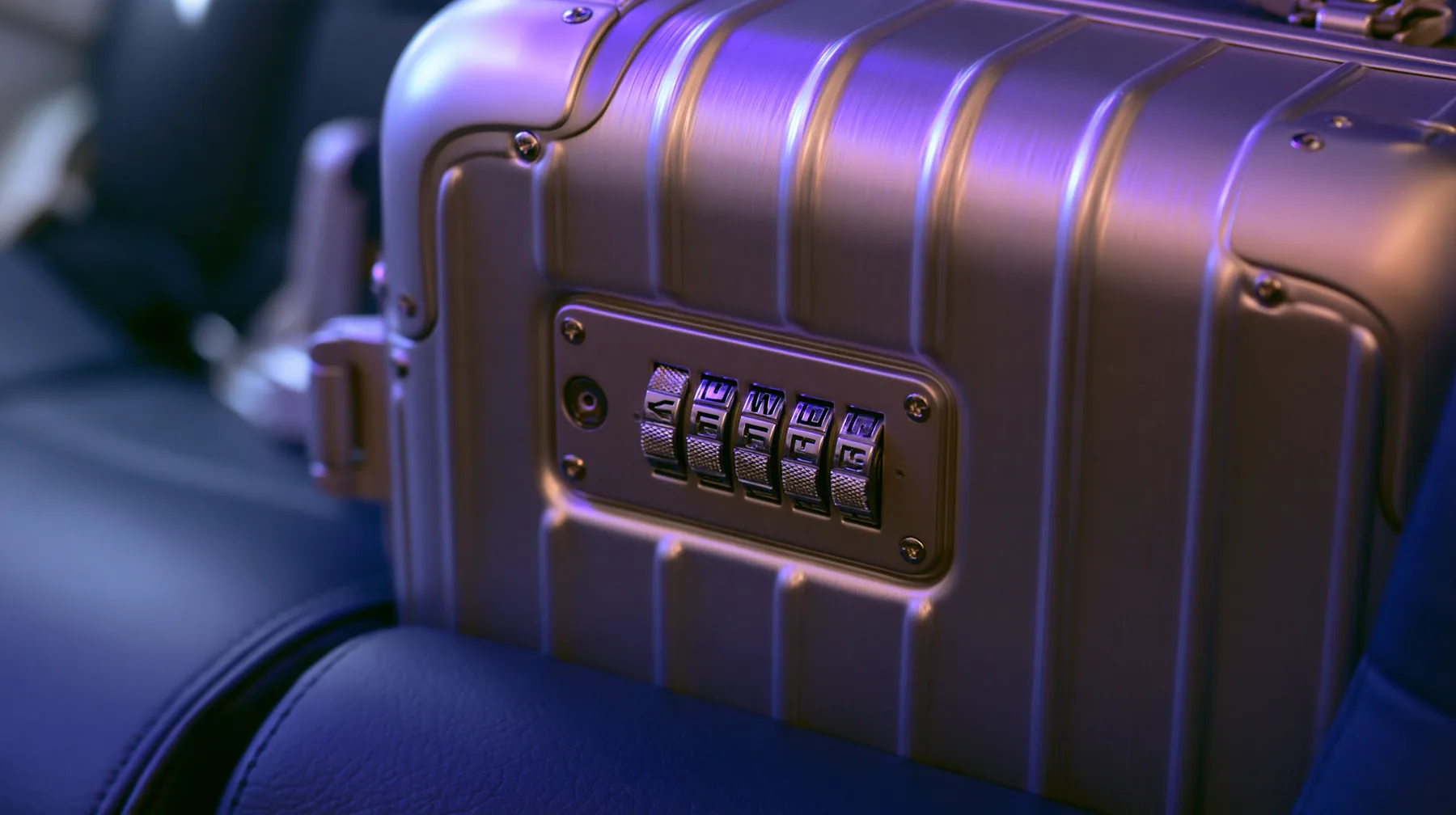 Jet-set editorial still life: a macro shot of a brushed-aluminum carry-on suitcase on a navy leather airplane seat, its built-in combination lock with five knurled metal letter tumbler wheels, violet cabin light…