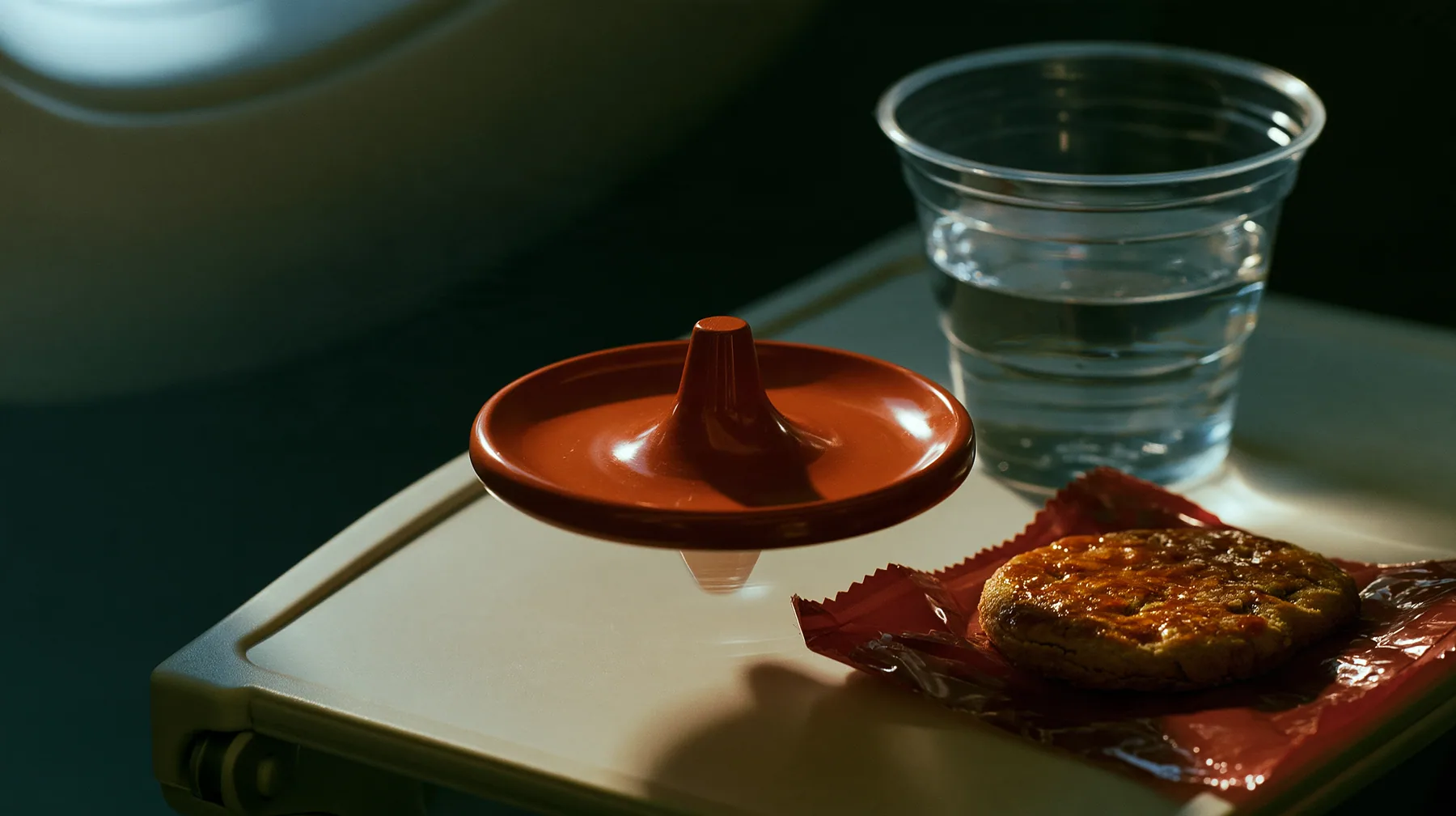 Jet-set editorial still life: a macro shot of an airplane tray table at night, a small polished clay-orange metal spinning top balanced on its point mid-spin, beside it a caramelized biscuit cookie in a red wrapper…