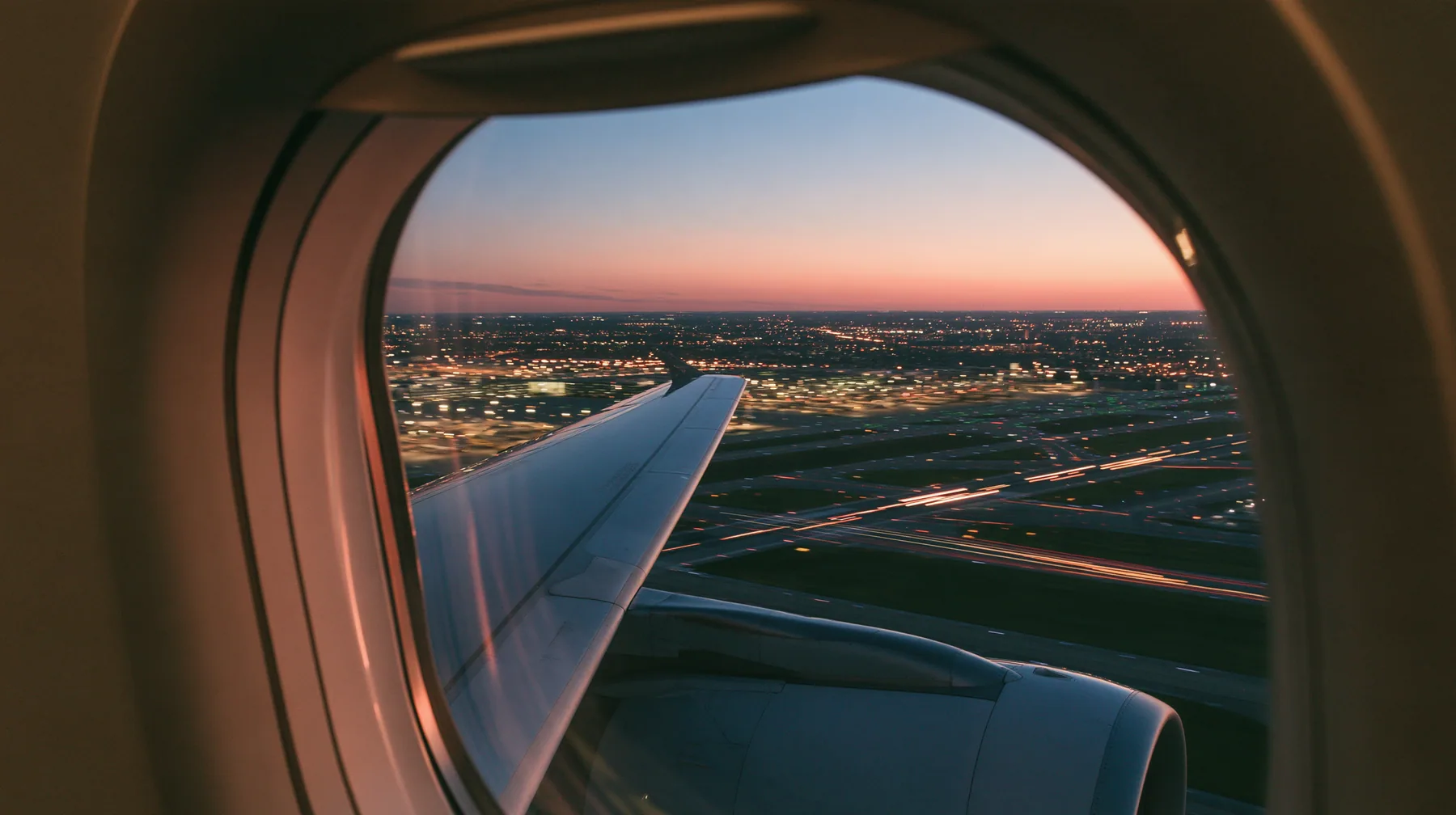 Jet-set editorial: the view out of an oval airliner window during takeoff at dusk, the wing and the engine below, the runway lights streaking past in long exposure, a pink and orange sky over the airport, the window…