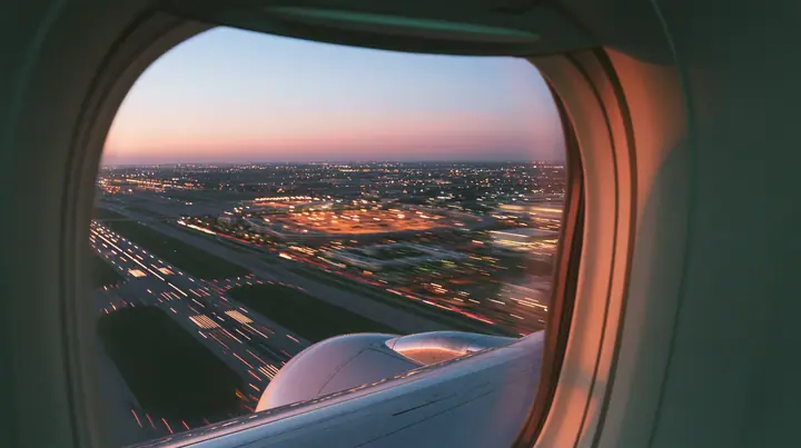 Jet-set editorial: the view out of an oval airliner window during takeoff at dusk, the wing and the engine below, the runway lights streaking past in long exposure, a pink and orange sky over the airport, the window…