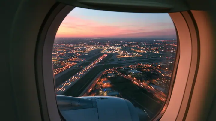 Jet-set editorial: the view out of an oval airliner window during takeoff at dusk, the wing and the engine below, the runway lights streaking past in long exposure, a pink and orange sky over the airport, the window…