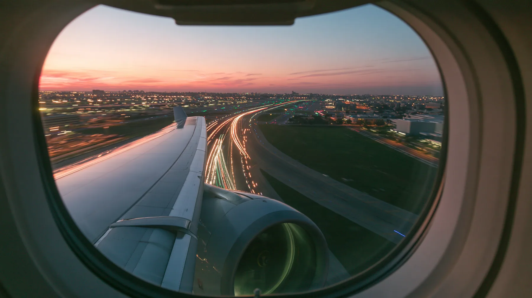 Jet-set editorial: the view out of an oval airliner window during takeoff at dusk, the wing and the engine below, the runway lights streaking past in long exposure, a pink and orange sky over the airport, the window…