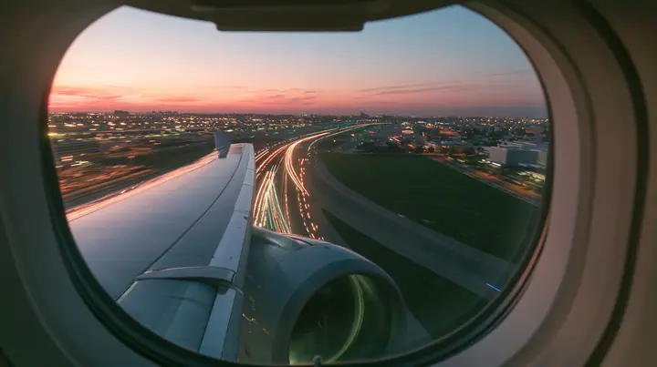 Jet-set editorial: the view out of an oval airliner window during takeoff at dusk, the wing and the engine below, the runway lights streaking past in long exposure, a pink and orange sky over the airport, the window…