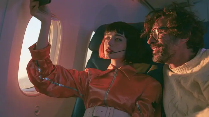 Jet-set high fashion editorial, a couture magazine story shot on a dawn flight: a medium shot across two airplane seats at dawn: a Caucasian American woman in her late twenties with pale skin and a glossy black blunt…