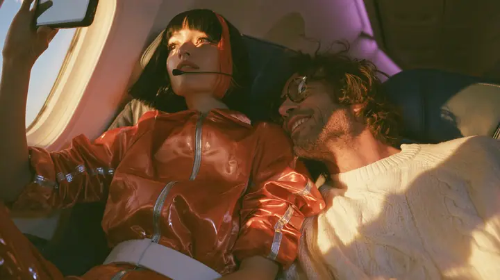 Jet-set high fashion editorial, a couture magazine story shot on a dawn flight: a medium shot across two airplane seats at dawn: a Caucasian American woman in her late twenties with pale skin and a glossy black blunt…