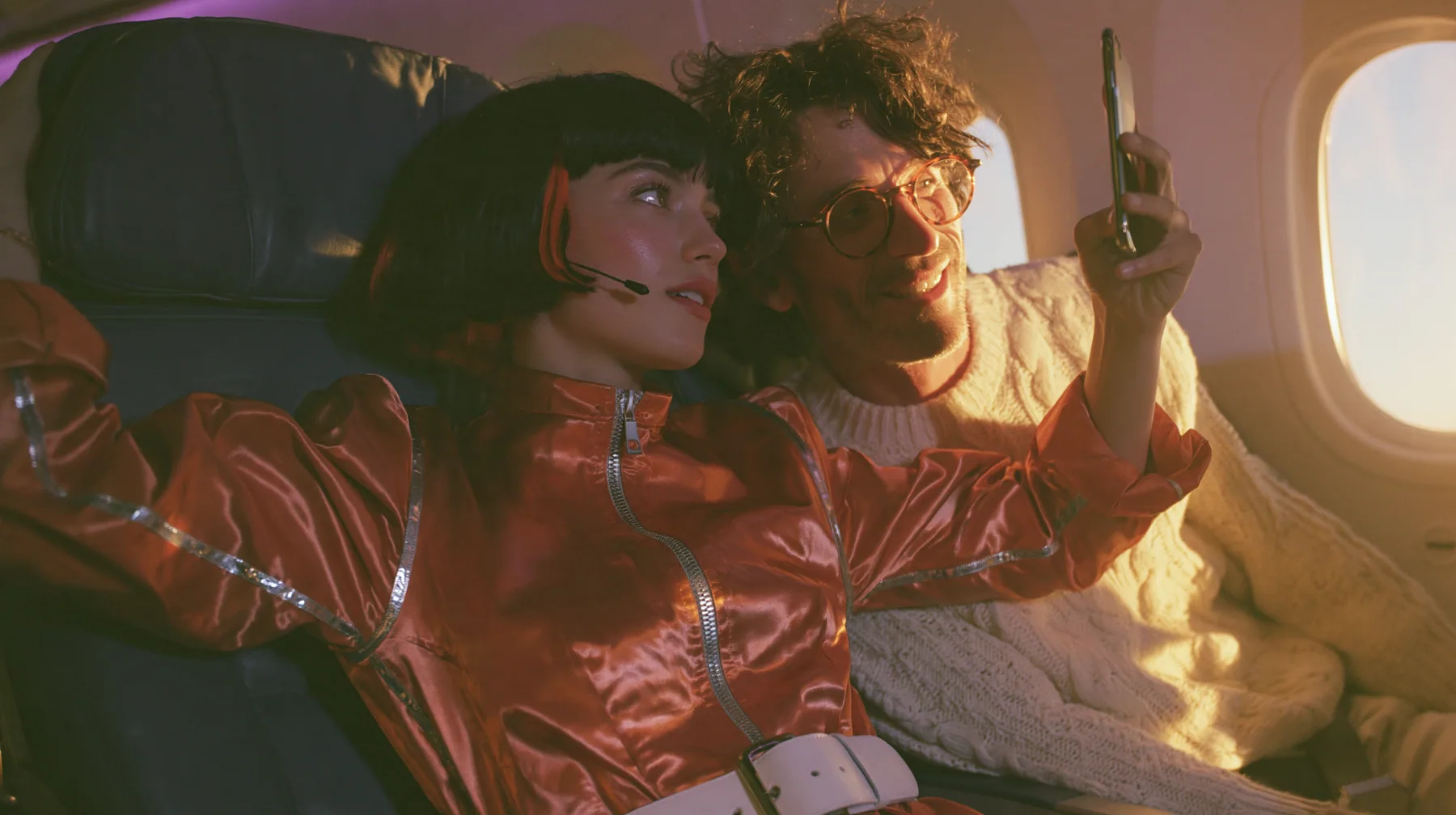Jet-set high fashion editorial, a couture magazine story shot on a dawn flight: a medium shot across two airplane seats at dawn: a Caucasian American woman in her late twenties with pale skin and a glossy black blunt…