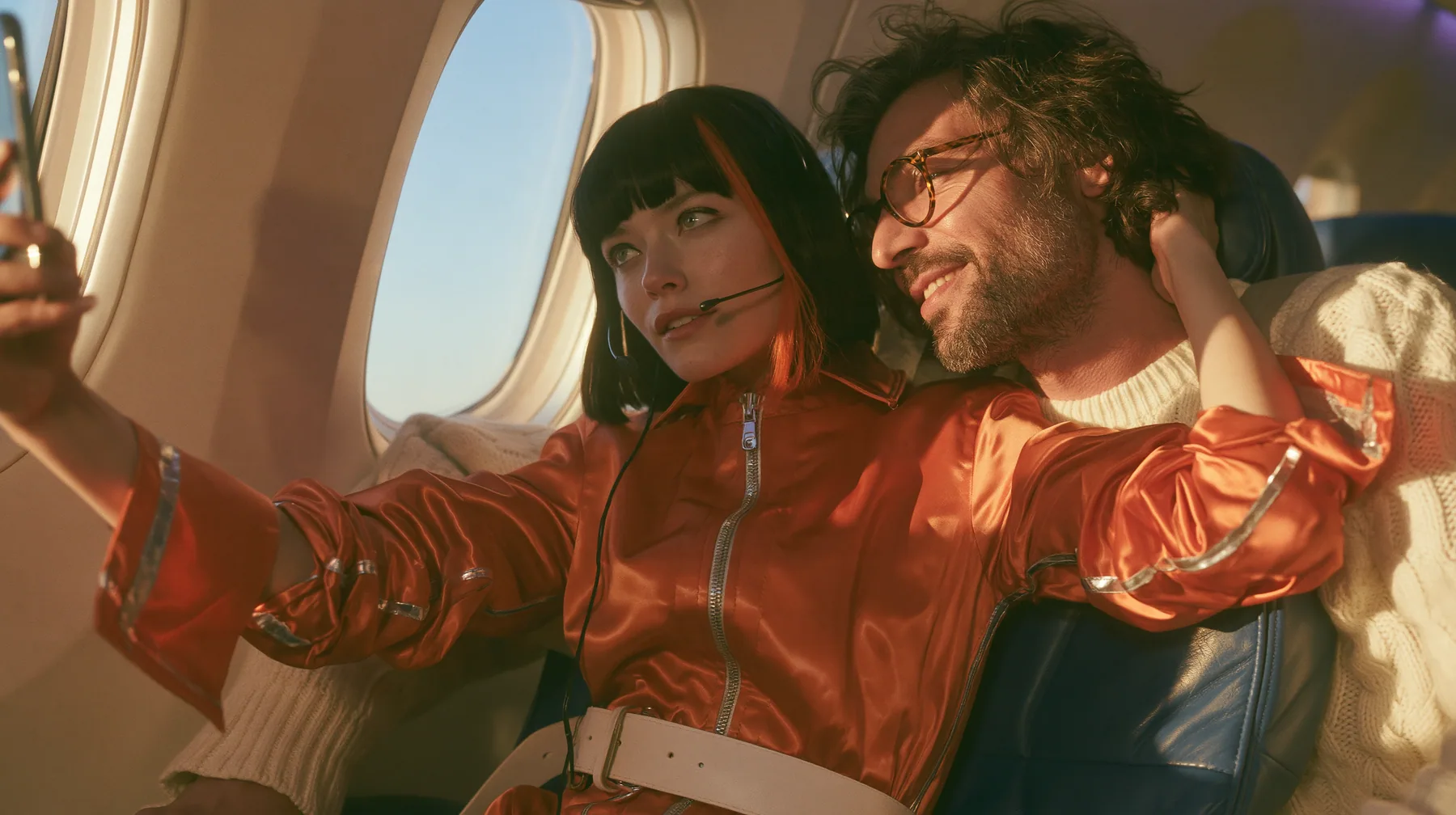 Jet-set high fashion editorial, a couture magazine story shot on a dawn flight: a medium shot across two airplane seats at dawn: a Caucasian American woman in her late twenties with pale skin and a glossy black blunt…