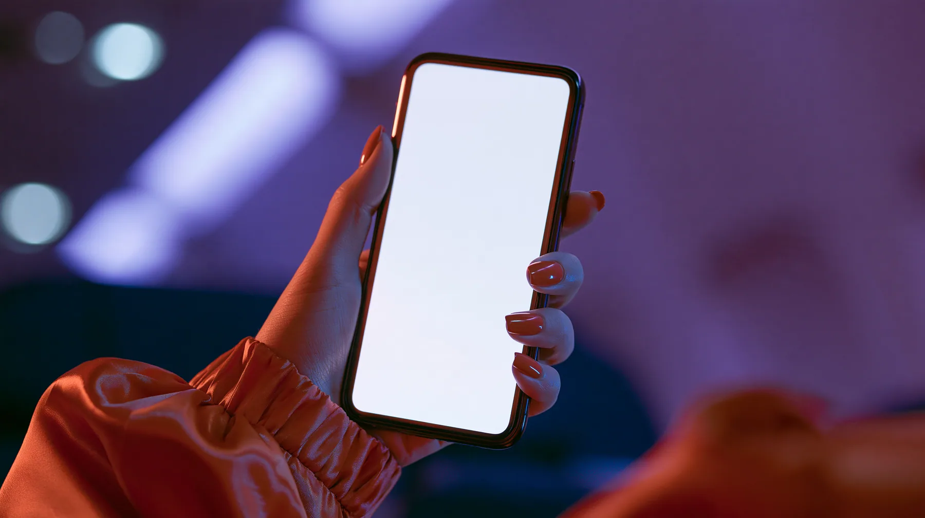 Jet-set editorial still life: an extreme close-up of a woman's hand with clay-orange nail polish holding a glossy black smartphone upright toward the lens, its screen a blank glowing white, the sleeve of a glossy…