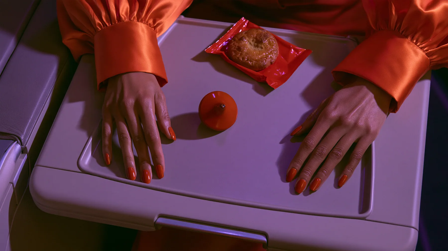 Jet-set editorial: a top-down overhead shot of an airplane tray table at night, a woman's hands with clay-orange nail polish and international-orange satin cuffs resting on its edges, a small polished clay-orange…