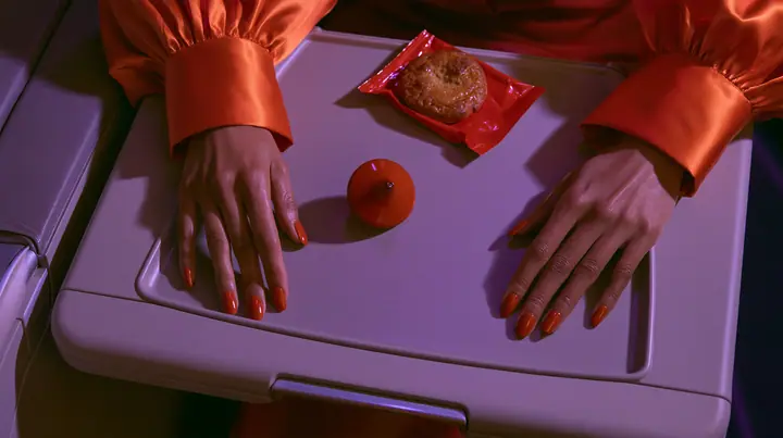 Jet-set editorial: a top-down overhead shot of an airplane tray table at night, a woman's hands with clay-orange nail polish and international-orange satin cuffs resting on its edges, a small polished clay-orange…