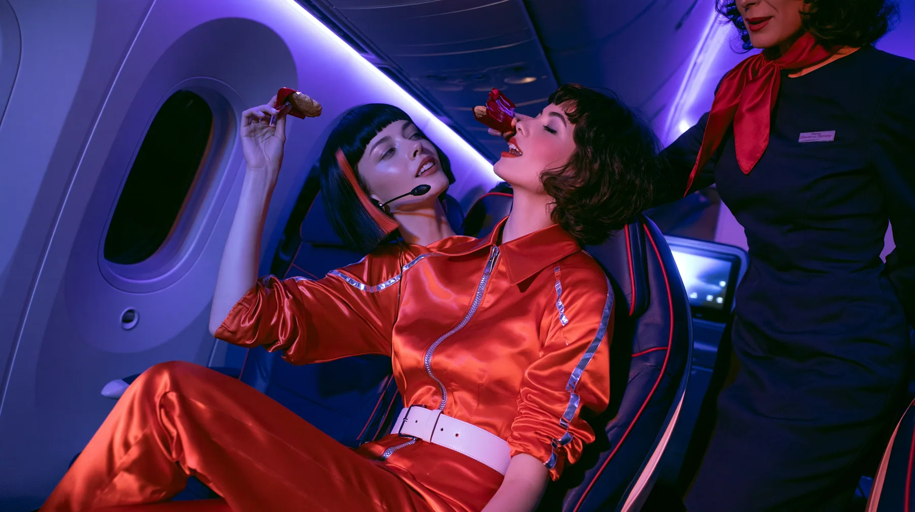 Jet-set high fashion editorial, a couture magazine story shot on a night flight: a medium two-shot in an airplane cabin: a Caucasian American woman in her late twenties with pale skin and a glossy black blunt bob…