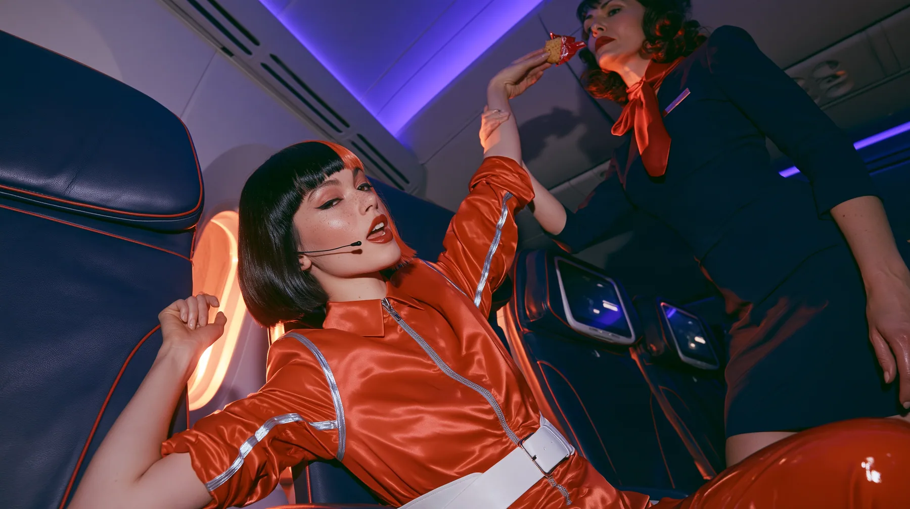 Jet-set high fashion editorial, a couture magazine story shot on a night flight: a medium two-shot in an airplane cabin: a Caucasian American woman in her late twenties with pale skin and a glossy black blunt bob…