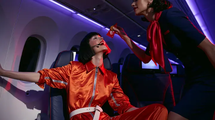 Jet-set high fashion editorial, a couture magazine story shot on a night flight: a medium two-shot in an airplane cabin: a Caucasian American woman in her late twenties with pale skin and a glossy black blunt bob…