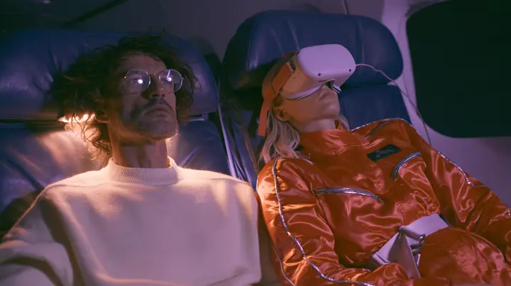 Jet-set fashion editorial, night flight: two airplane seats at night: a man in his forties with tousled hair, round glasses and a cream sweater sits wide awake under his reading light staring sideways; beside him a…