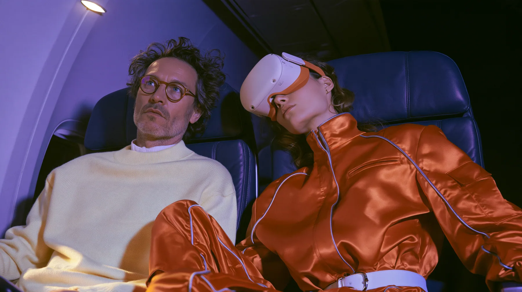 Jet-set fashion editorial, night flight: two airplane seats at night: a man in his forties with tousled hair, round glasses and a cream sweater sits wide awake under his reading light staring sideways; beside him a…