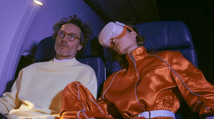 Jet-set fashion editorial, night flight: two airplane seats at night: a man in his forties with tousled hair, round glasses and a cream sweater sits wide awake under his reading light staring sideways; beside him a…