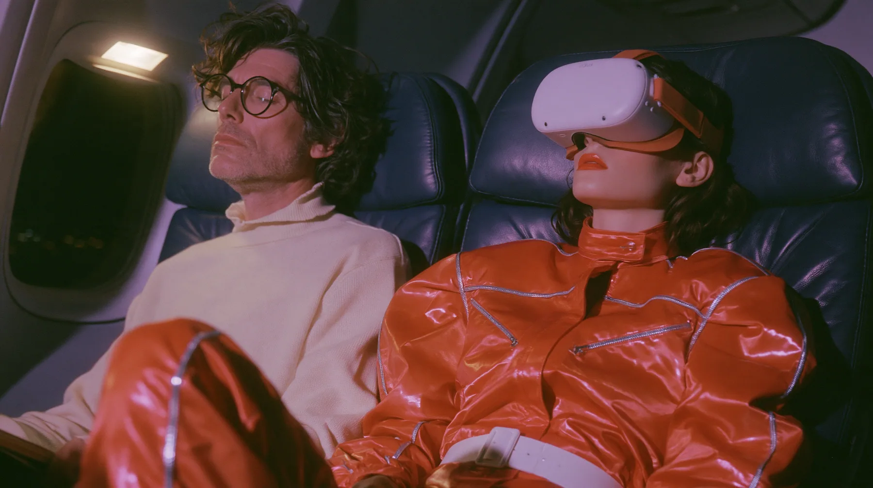 Jet-set fashion editorial, night flight: two airplane seats at night: a man in his forties with tousled hair, round glasses and a cream sweater sits wide awake under his reading light staring sideways; beside him a…