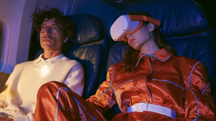 Jet-set fashion editorial, night flight: two airplane seats at night: a man in his forties with tousled hair, round glasses and a cream sweater sits wide awake under his reading light staring sideways; beside him a…