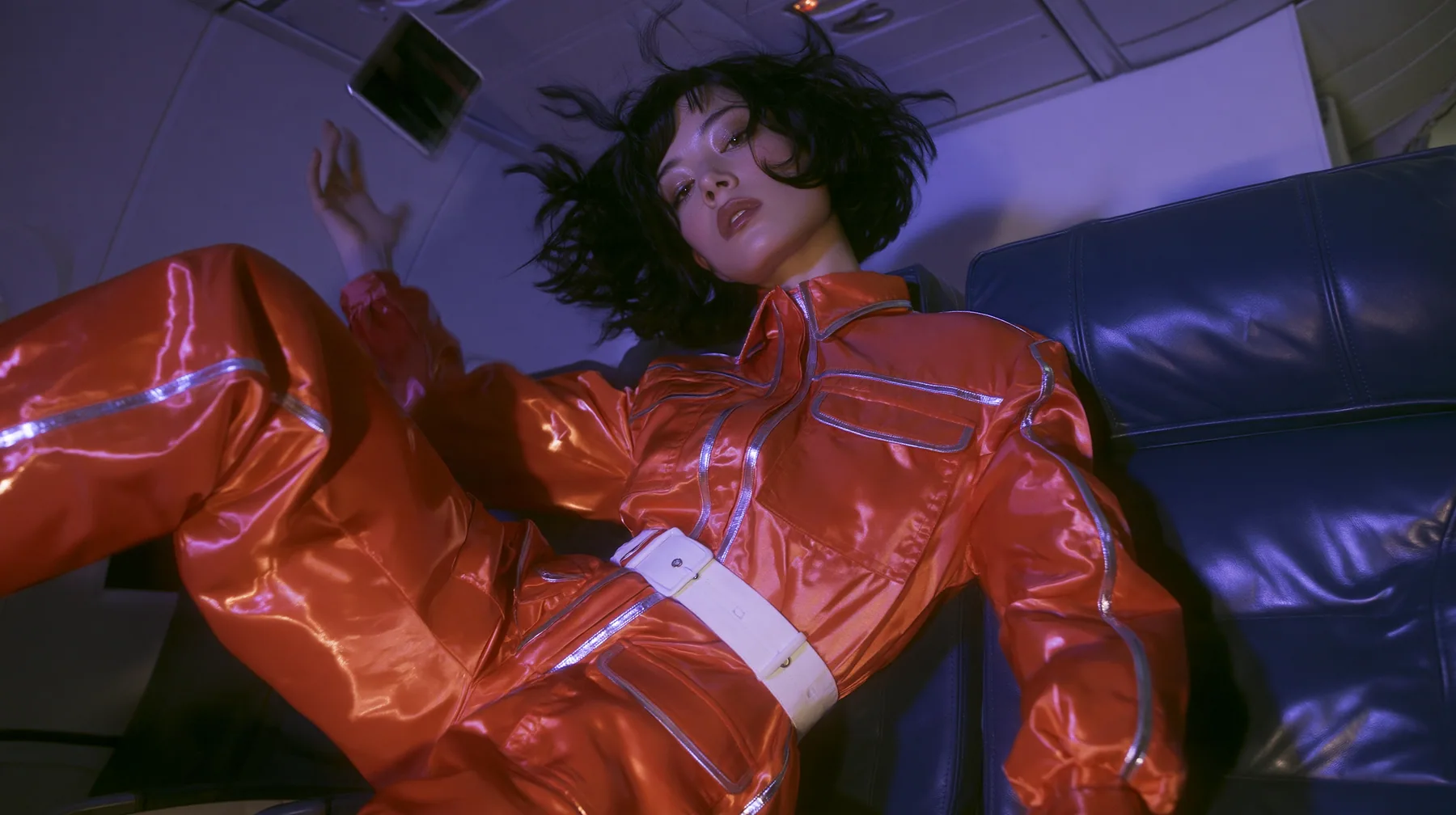 Jet-set fashion editorial, night flight: medium shot of a woman in a glossy international-orange satin flight jumpsuit with silver piping and a white belt with a floating black bob, weightless in an airliner cabin,…