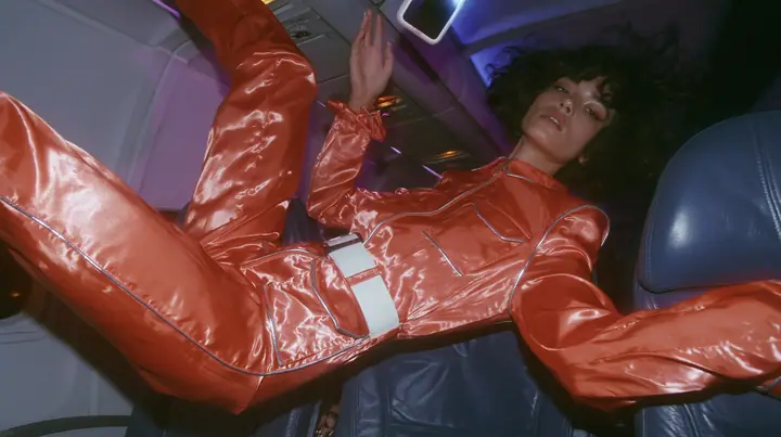 Jet-set fashion editorial, night flight: medium shot of a woman in a glossy international-orange satin flight jumpsuit with silver piping and a white belt with a floating black bob, weightless in an airliner cabin,…