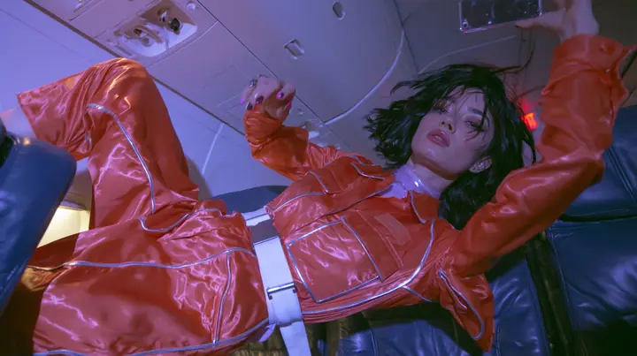 Jet-set fashion editorial, night flight: medium shot of a woman in a glossy international-orange satin flight jumpsuit with silver piping and a white belt with a floating black bob, weightless in an airliner cabin,…