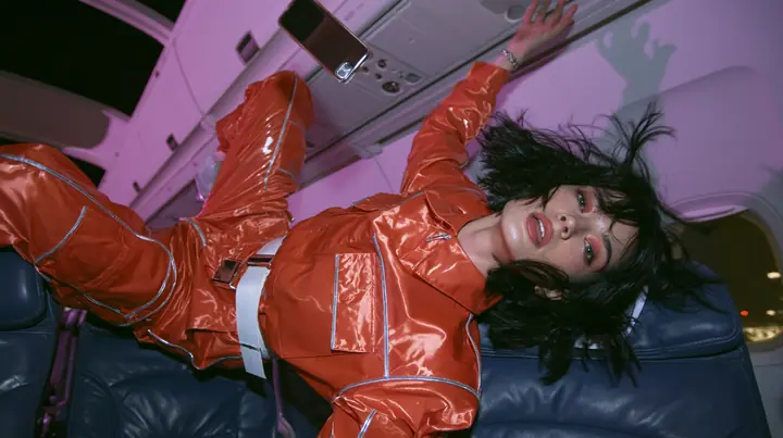 Jet-set fashion editorial, night flight: medium shot of a woman in a glossy international-orange satin flight jumpsuit with silver piping and a white belt with a floating black bob, weightless in an airliner cabin,…