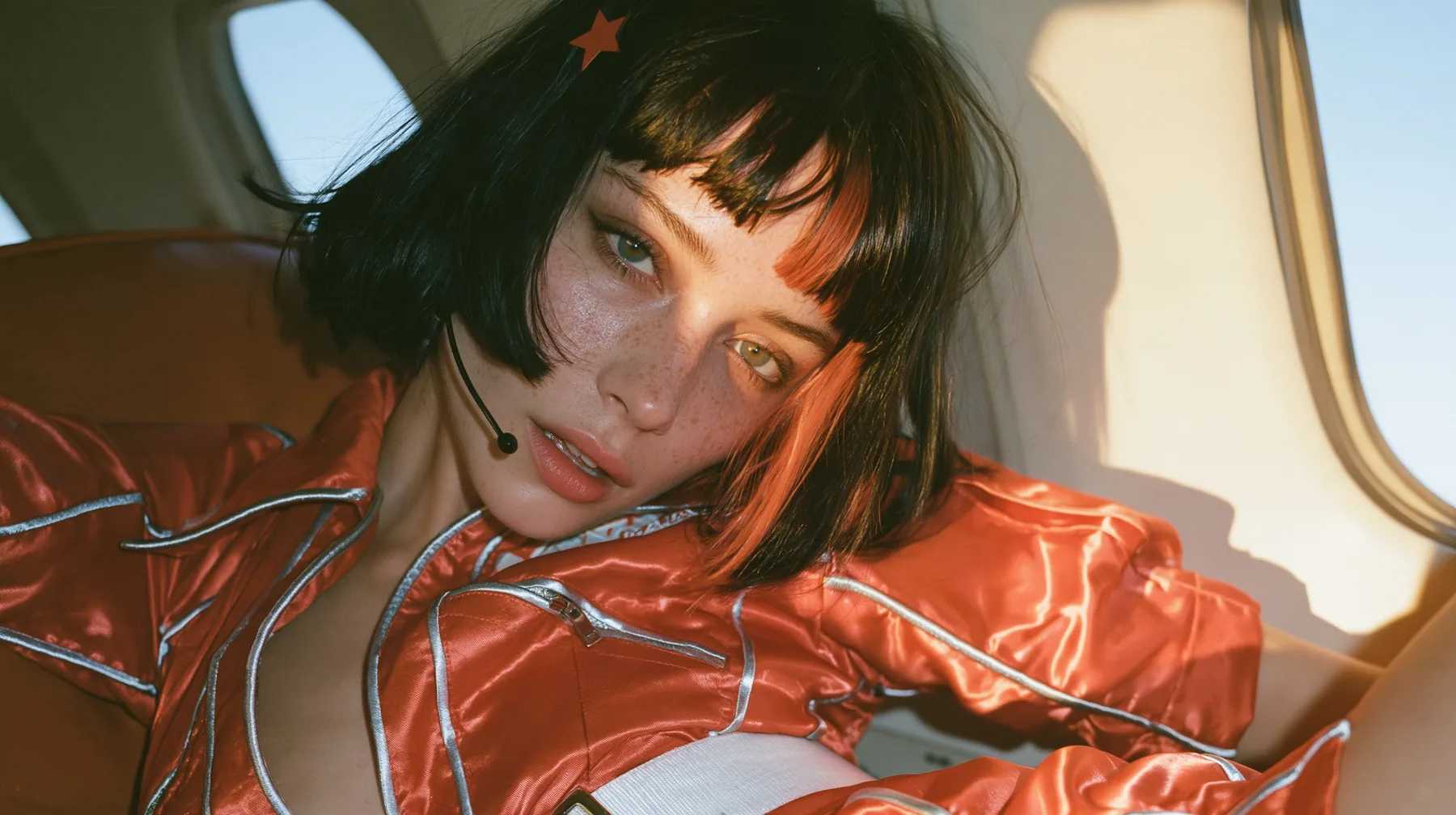 Jet-set fashion editorial, dawn flight: medium close-up of a Caucasian American woman in her late twenties with a grown-up angular face, pale freckled skin, a glossy black blunt bob with heavy bangs and one…