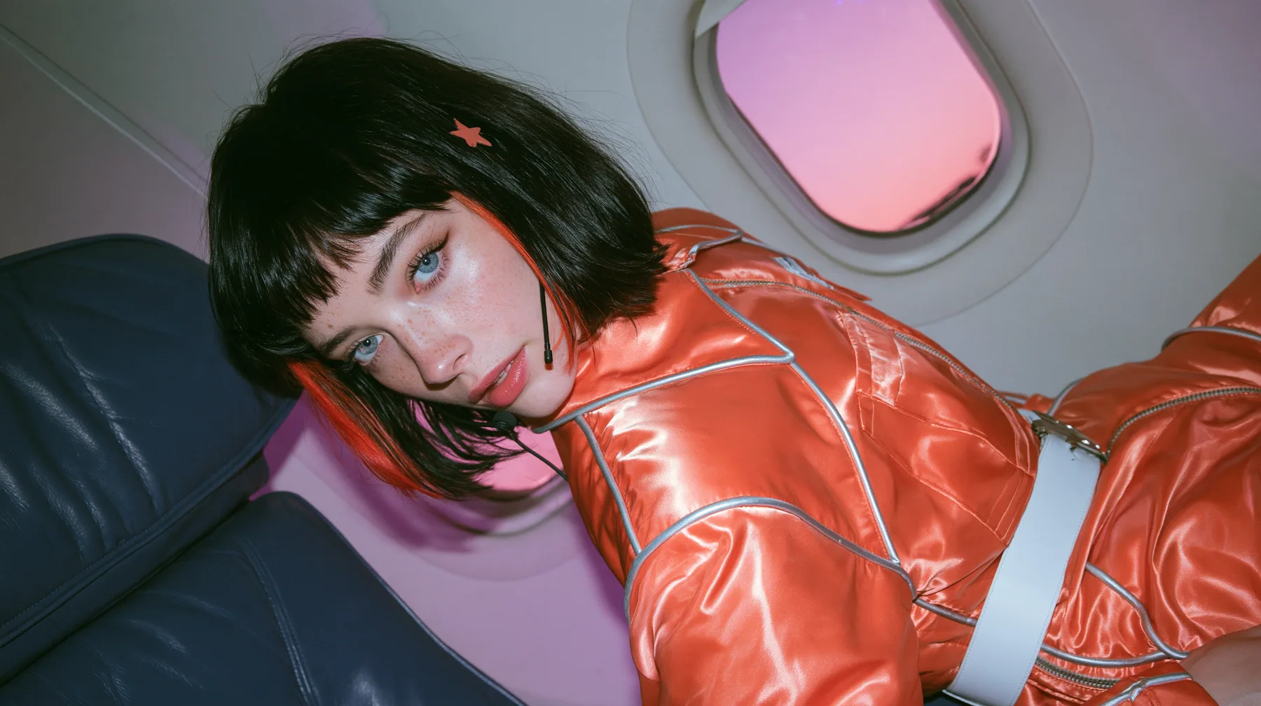 Jet-set fashion editorial, night flight: medium close-up of a Caucasian American woman in her late twenties with a grown-up angular face, pale freckled skin, a glossy black blunt bob with heavy bangs and one…