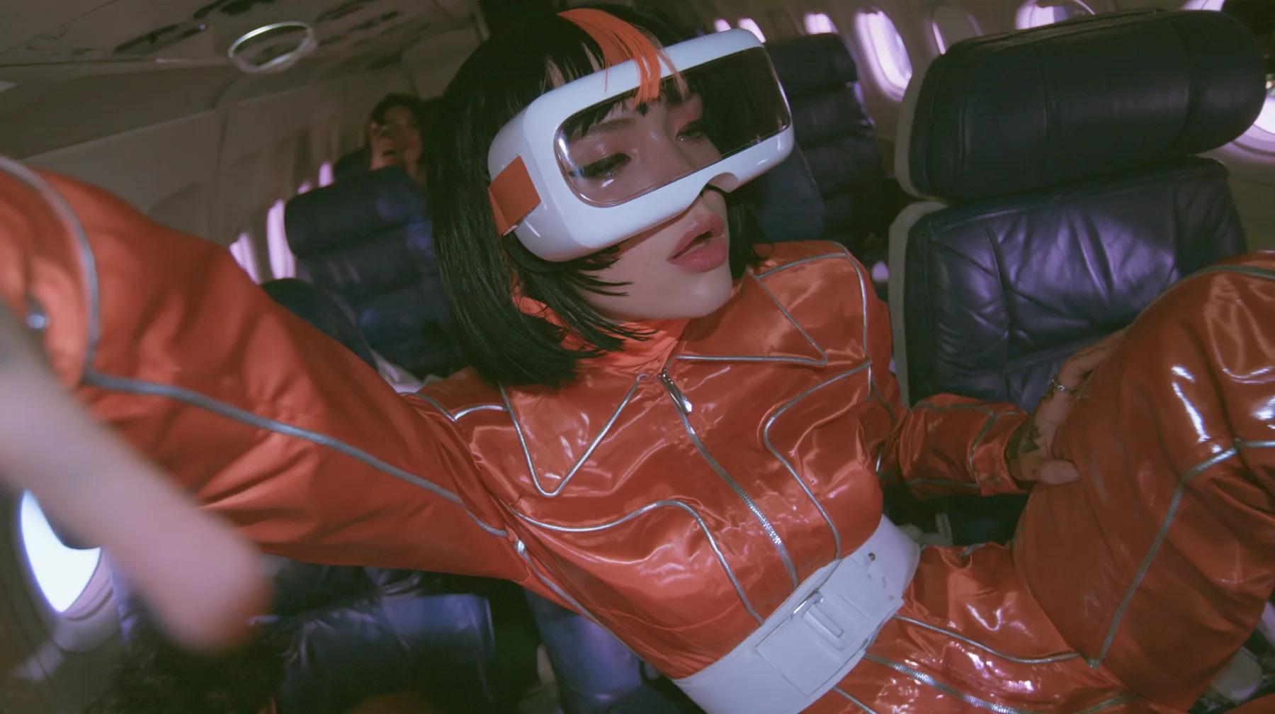 Jet-set fashion editorial, night flight: medium close-up of a woman in a glossy international-orange satin flight jumpsuit with silver piping and a white belt with a black bob and one orange streak, a glossy white VR…