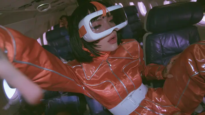Jet-set fashion editorial, night flight: medium close-up of a woman in a glossy international-orange satin flight jumpsuit with silver piping and a white belt with a black bob and one orange streak, a glossy white VR…