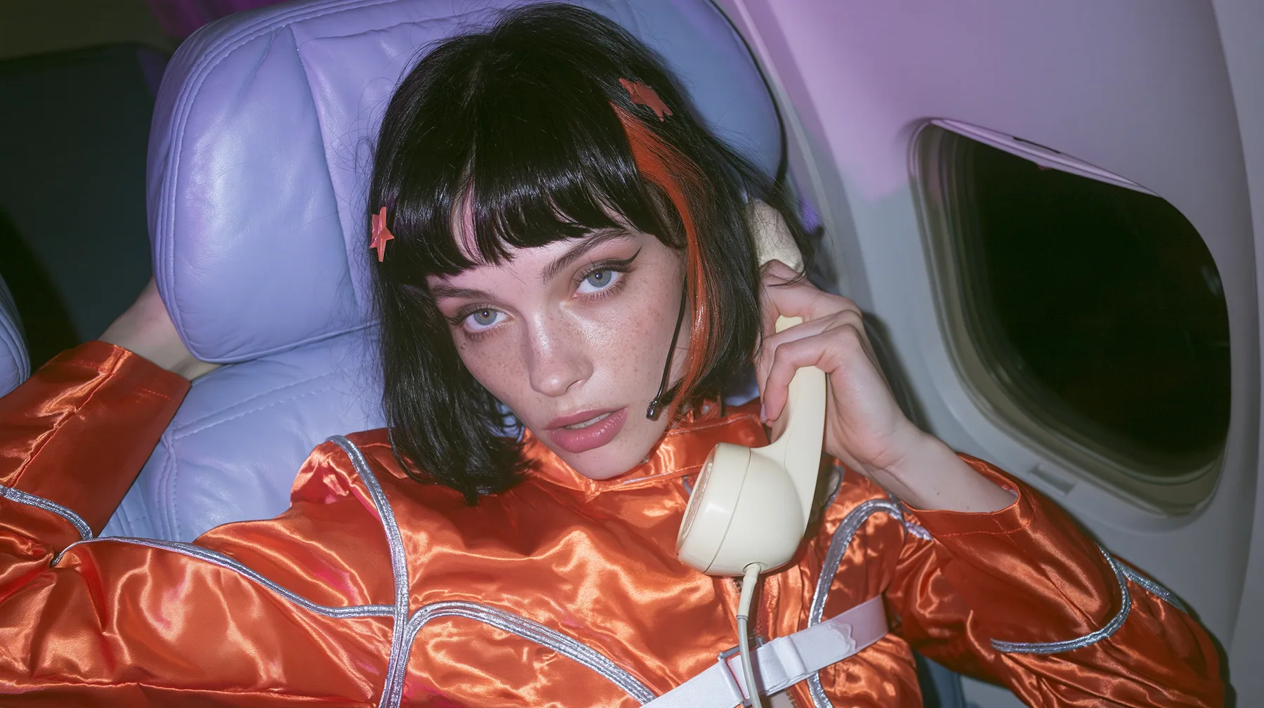 Jet-set fashion editorial, night flight: an eye-level medium shot of a Caucasian American woman in her early thirties, a grown woman with elegant mature features and defined cheekbones, pale freckled skin, a glossy…