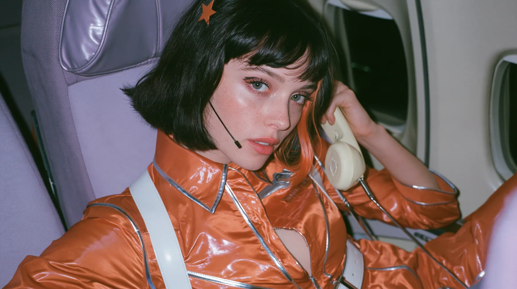 Jet-set fashion editorial, night flight: an eye-level medium shot of a Caucasian American woman in her early thirties, a grown woman with elegant mature features and defined cheekbones, pale freckled skin, a glossy…