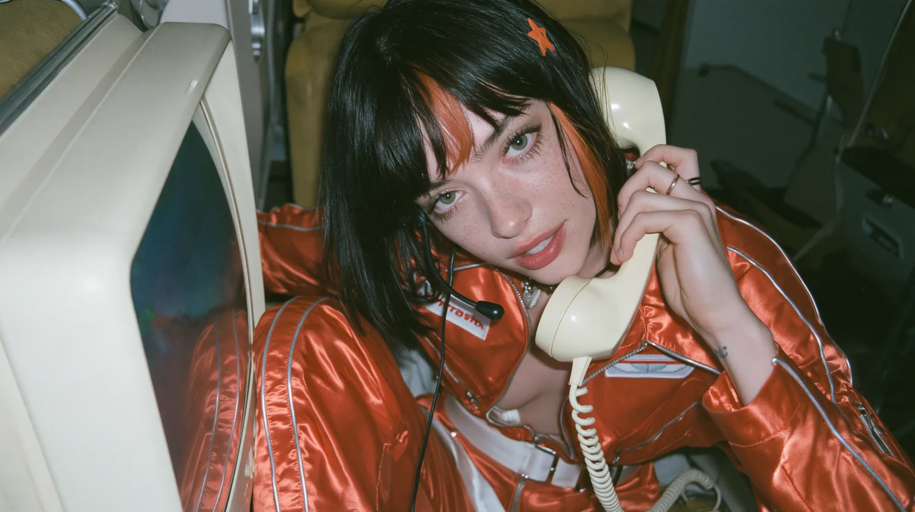 Jet-set fashion editorial, night flight: an eye-level medium shot of a Caucasian American woman in her early thirties, a grown woman with elegant mature features and defined cheekbones, pale freckled skin, a glossy…
